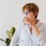 Focused female in casual shirt and eyeglasses drinking pure water from glass while standing against white wall and green houseplants