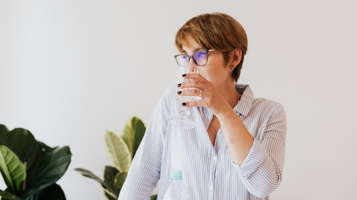 Focused female in casual shirt and eyeglasses drinking pure water from glass while standing against white wall and green houseplants